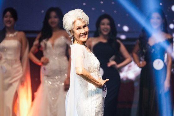 Choi Soon-hwa smiles on stage in front of other contestants, wearing a white dress