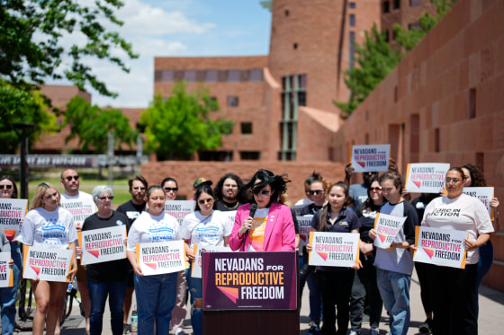Lindsey Harmon speaks outside behind a podium while people stand on either side of her holding signs that say "Nevadans for Reproductive Freedom"