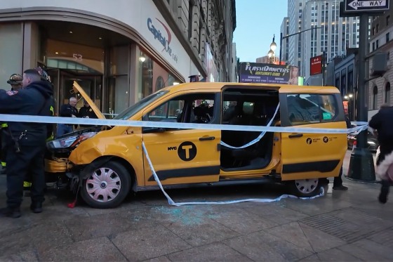 A crashed taxi van sits atop a sidewalk as it's cordoned off by caution tape and surrounded by first responders.