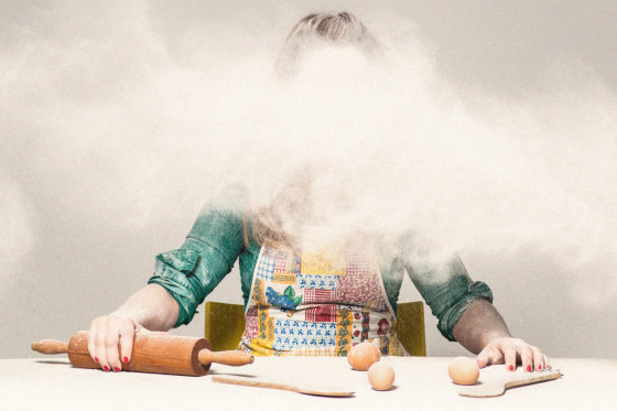 A woman in an apron, face obscured by a cloud of flour, sits at a table with a rolling pin and eggs