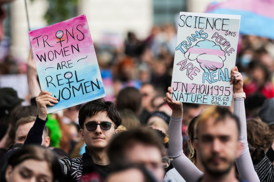 Two people hold signs outside in a crowd, sign on the left says "Trans women are real women", sign on the right says "Science says trans people are real"