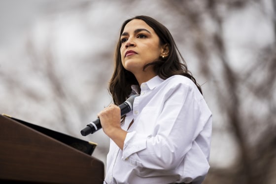 Alexandra Ocasio-Cortez holds a microphone to her chest while standing at a podium outside