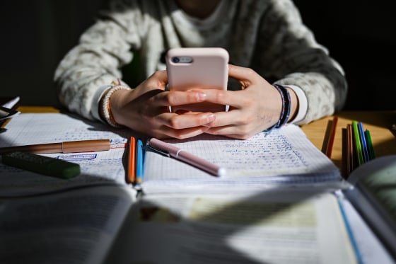 A child's hands hold a cellphone on top of a desk with homework and pens
