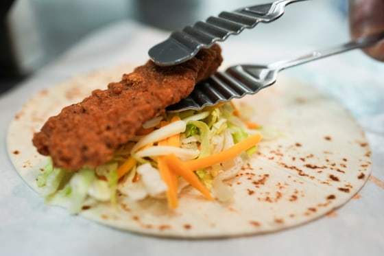 A pair of tongs places a chicken tender onto a tortilla with lettuce and shredded cheese
