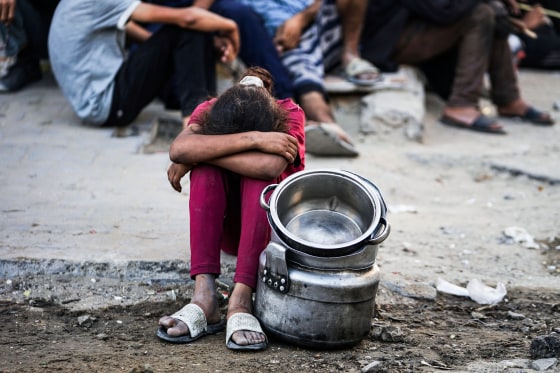 A girl sits on the ground with her head tucked into her legs, empty pots sit beside her