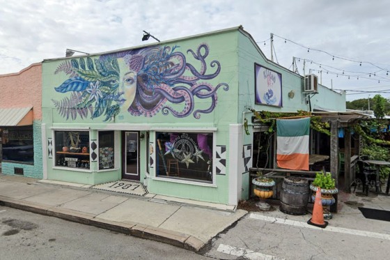 The exterior of Kilted Mermaid bar, a mint green painted building with a painting of a woman's face with plants and tentacles reaching out from either side
