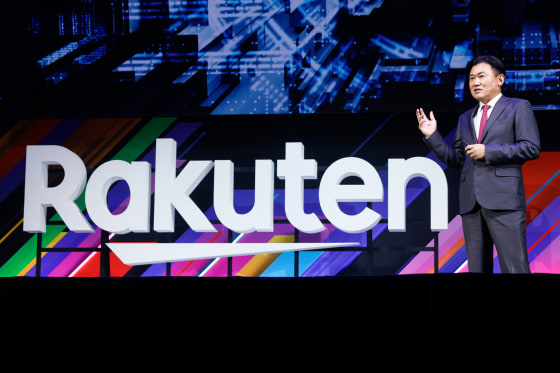 Hiroshi Mikitani speaks on stage next to large white text that reads "Rakuten"