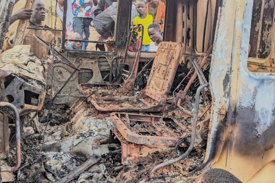 People gather around the charred remains of a burned vehicle