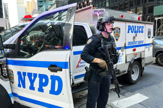 An NYPD officer carrying a gun stands next to an NYPD emergency service vehicle parked on the street outside