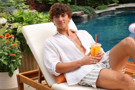 Gavin Casalegno sits in a pool chair by the pool holding a Dunkin Donuts Golden Hour refresher drink