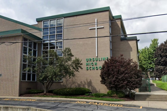 The exterior of Ursuline High School, a large white cross hangs on the beige brick side of the building on top of green text that reads "Ursuline High School"