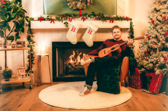 Hunter Hayes sits on chair in a living room holding a guitar, with the fireplace going and Christmas tree lights on