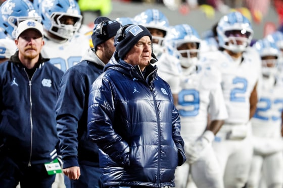 Bill Belichick stands on the sideline with players outside on the football field