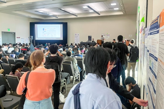 People stand and sit inside of a conference room, with a large projector screen showing a slideshow