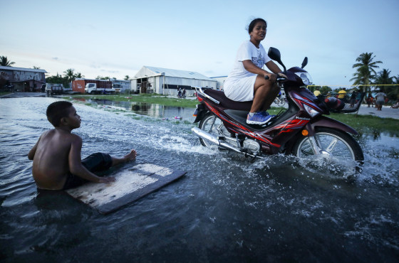A woman rides her scooter through floodwaters next to a boy sitting near a plank on the flooded street