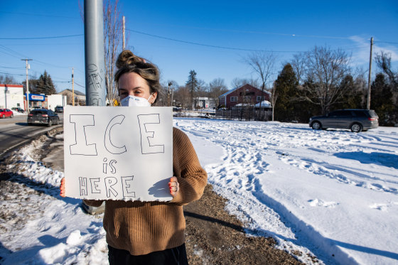An activist holds a sign that reads "ICE IS HERE" near an alleged federal agent vehicle.