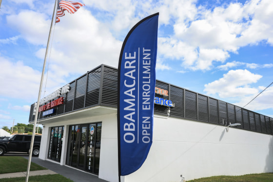 A blue sign reading OBAMACARE OPEN ENROLLMENT is displayed outside a white one-story building.