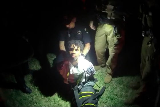 A young black man is held from behind as he kneels in the grass, illuminated by flashlights.