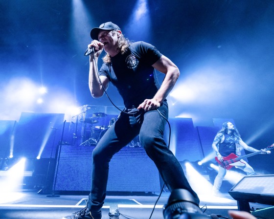 Brad Arnold holds a microphone while singing onstage under blue lights. He's wearing a black t-shirt and black baseball cap.