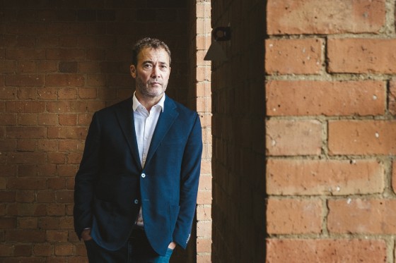 Will Lewis leans against a brick wall wearing a navy blazer and white shirt without a necktie.