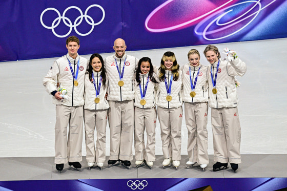 Seven olympic figure skaters in white warmup suits stand with gold medals around their necks.