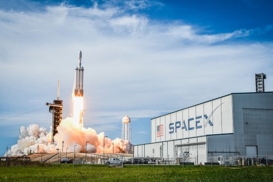 A rocket launches off a pad next to a building with large text on the front that reads "SpaceX"