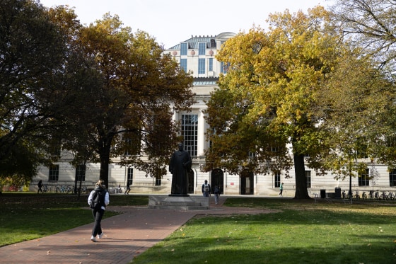 A student wearing a backpack walks away and toward a large building surrounded by trees.