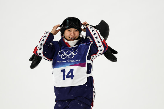 Chloe Kim stands in the snow and smiles as she removes ski goggles from her head.