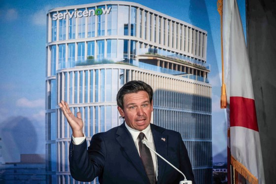 Ron DeSantis gestures with an open hand in front of a backdrop depicting a high rise office building.