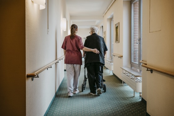 Rear view of female caregiver assisting senior woman walking with mobility walker in corridor