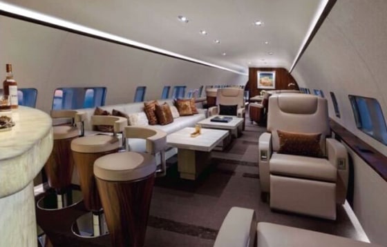 The interior of a plane, beige reclining seats and bar stools can be seen