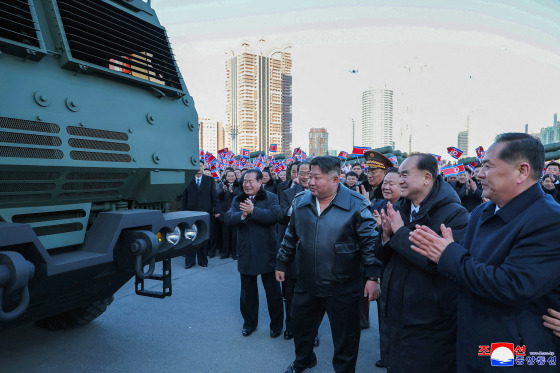 North Korean leader Kim Jong Un  amongst a group of men while looking at appears to be a military truck. People stands behind holding North Korea's flag.