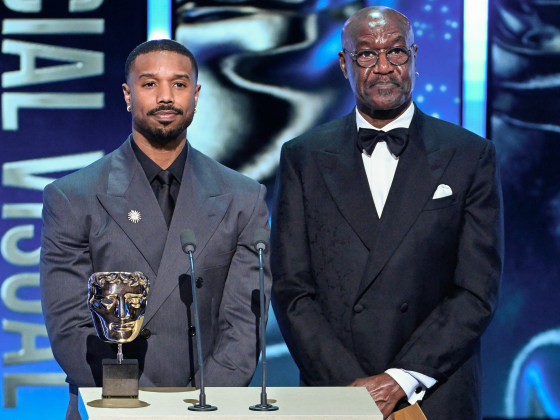 Michael B. Jordan and Delroy Lindo stand at a lectern on stage during an award ceremony. They look nonplussed.