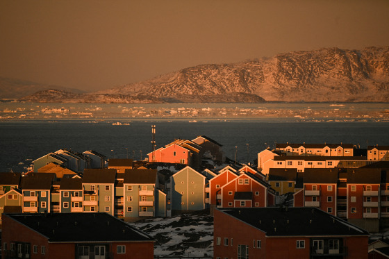 Orange sunset light illuminates three-story buildings on a shoreline. Ice floats it the water offshore. 