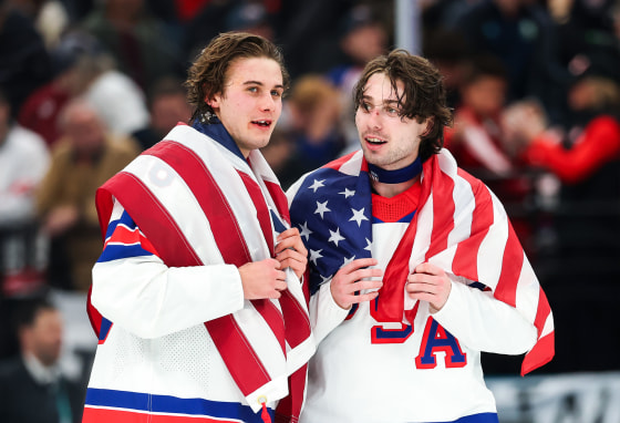 Jack Hughes, left, and Quinn Hughes speak with American flags draped over their shoulders on the ice