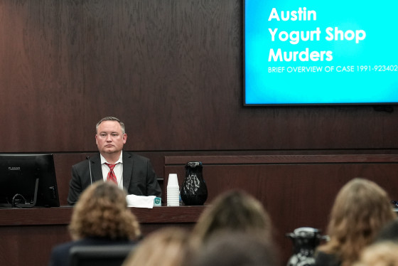 A man wearing a red necktie and black suit jacket is seated at a table during a court hearing. A large projector screen reads "Austin Yogurt Shop Murders".