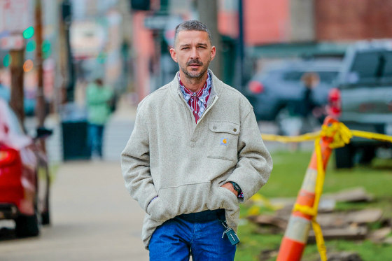 Shia LaBeouf walks on a sidewalk with both hands in the pocket of his sweatshirt.