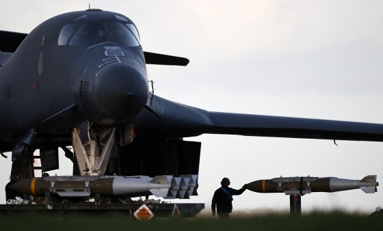 A soldier wearing earmuffs with one hand on a large missile is silhouetted against the sky.