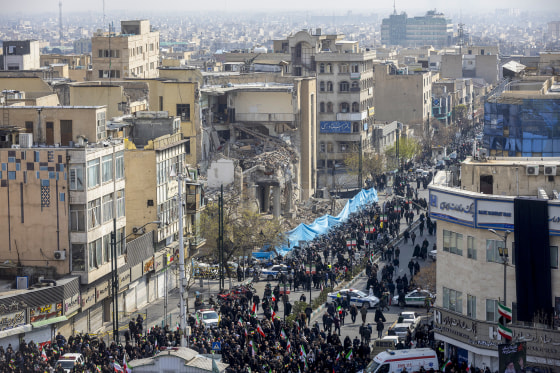 Funeral held with damaged buildings in the background.