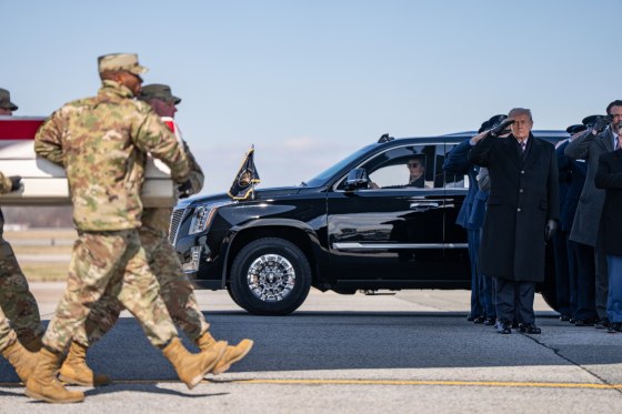 Army members carry a casket across an aircraft tarmac as Donald Trump and company stand to watch while saluting