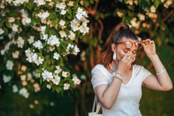 Summer time. unhappy elegant female in white shirt with handkerchief and eyeglasses has an allergy attack near flowering tree.