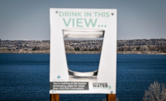 A sign with a water glass on it appears in soft focus in front of a large mountain lake. 