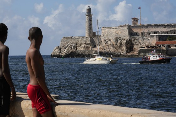 Two shirtless young men watch a tugboat pulling a sailboat from the shore.