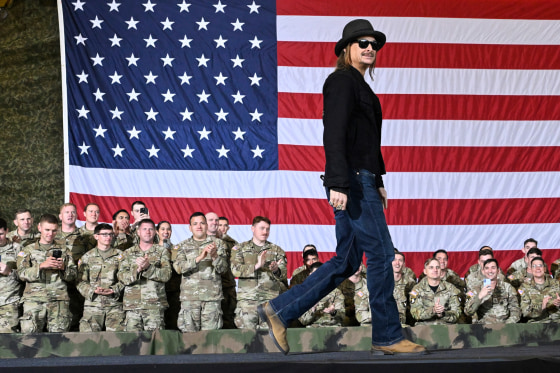 Kid Rock walks across a stage, military in the audience stand to clap, an American flag backdrop is behind them all
