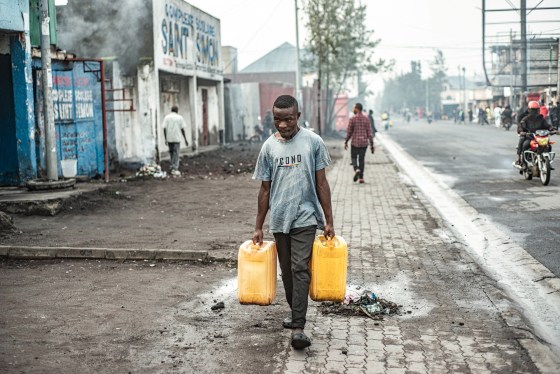 A man carries two yellow water jugs outside on the sidewalk