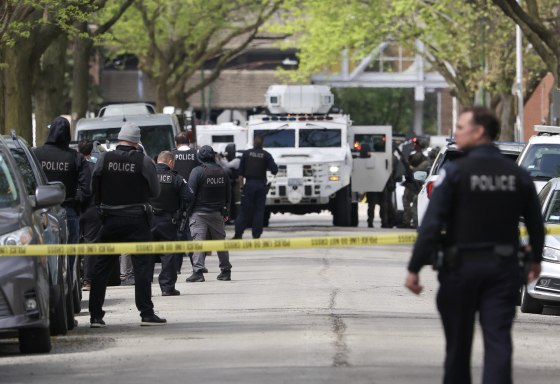 Several police officers in black flak jackets stand behind yellow tape in a city street.
