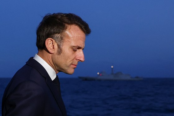 Emmanuel Macron appears in profile view in front of a battleship floating in the sea at dusk.