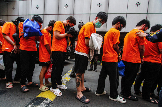 People wearing orange t-shirts with orange zip ties binding their wrists are walked by police in a line