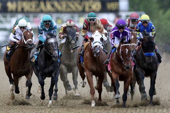 A tight group of racehorses and jockeys race toward the camera on a dirt track.