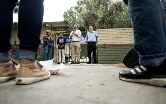 Three politicians stand in a row against a brick wall, seen from ground level between two people's legs.